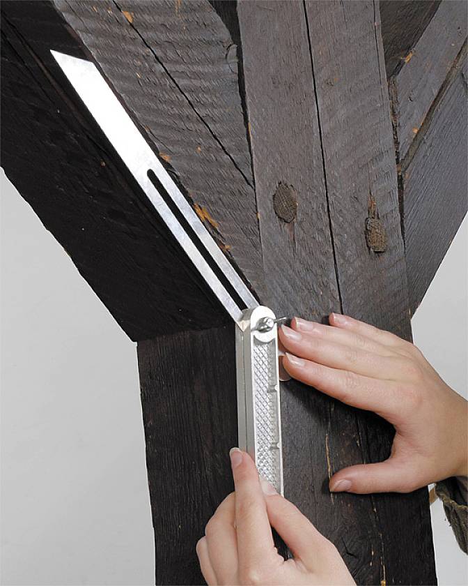 Osoba używa kątomierza do zmierzenia kąta drewnianego belki. Belka ma ciemną powierzchnię drewna.