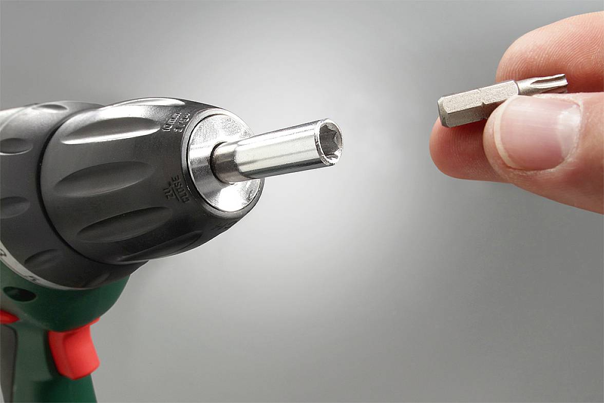 Jedna ręka trzyma końcówkę wkrętaka przed wiertarką. Wiertarka jest czarno-zielona, z uchwytem w kolorach czerwono-czarnym.
