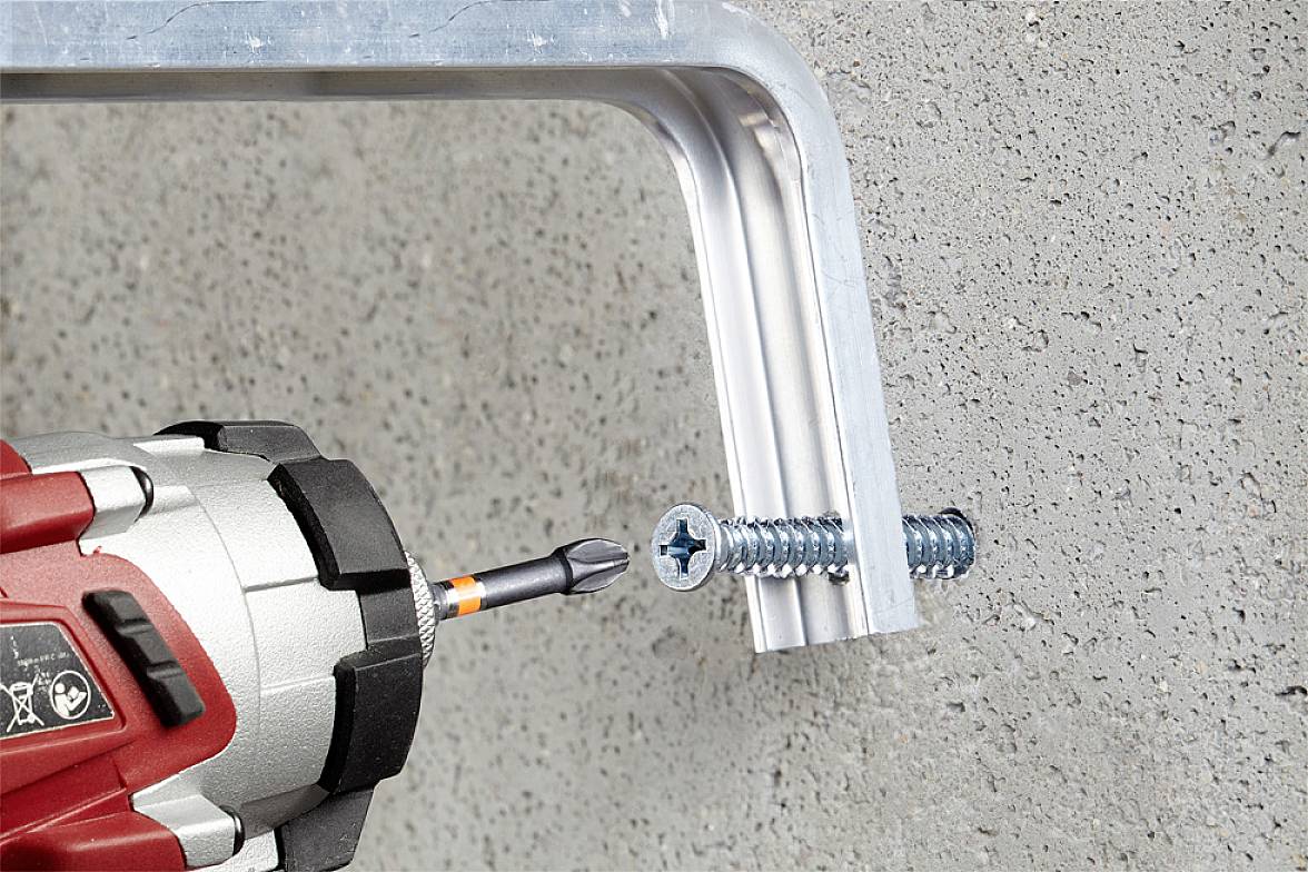 Wkrętak akumulatorowy mocuje metalowy element do szarej ściany betonowej za pomocą srebrnej końcówki śrubowej.