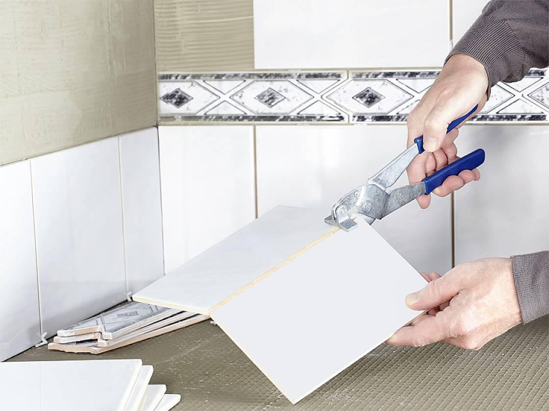 Osoba ucina białą płytkę szczypcami do płytek w kuchni z wyłożoną płytkami ścianą.