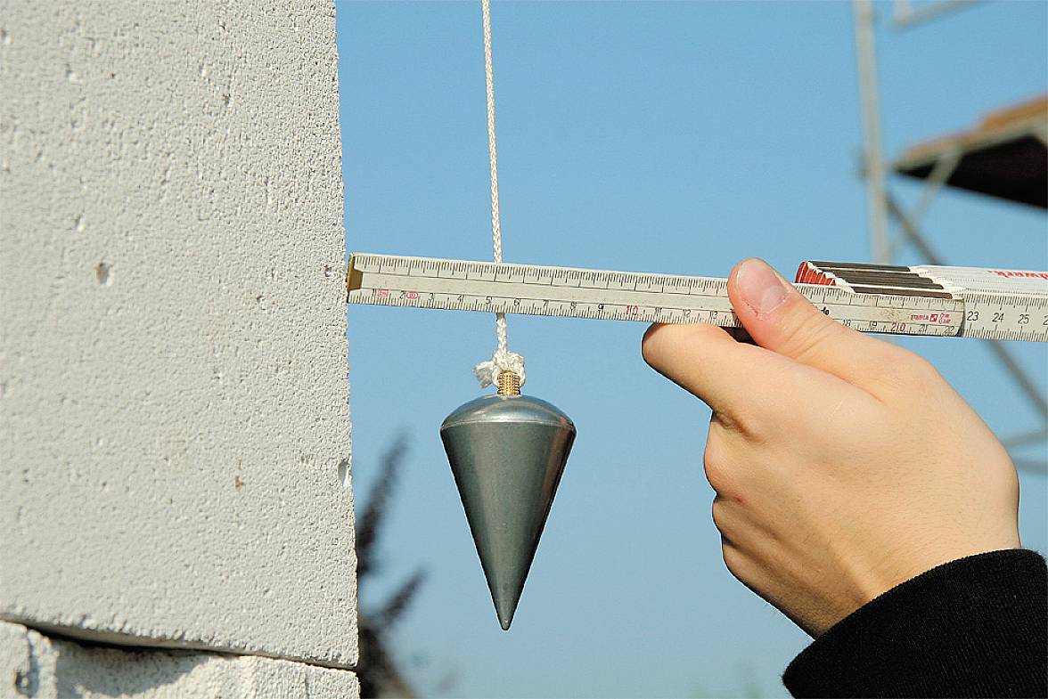 Jedna ręka trzyma miarkę, która za pomocą pionu sprawdza pionowość ściany.