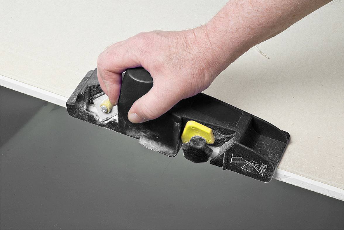 Dłoń trzyma czarne narzędzie, które ścina krawędź płyty gipsowo-kartonowej.