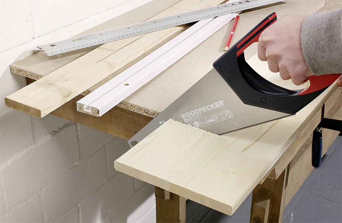 'Ramię trzyma piłę ręczną i przecina deskę drewnianą na stole warsztatowym, obok linijek i czerwonego ołówka.'