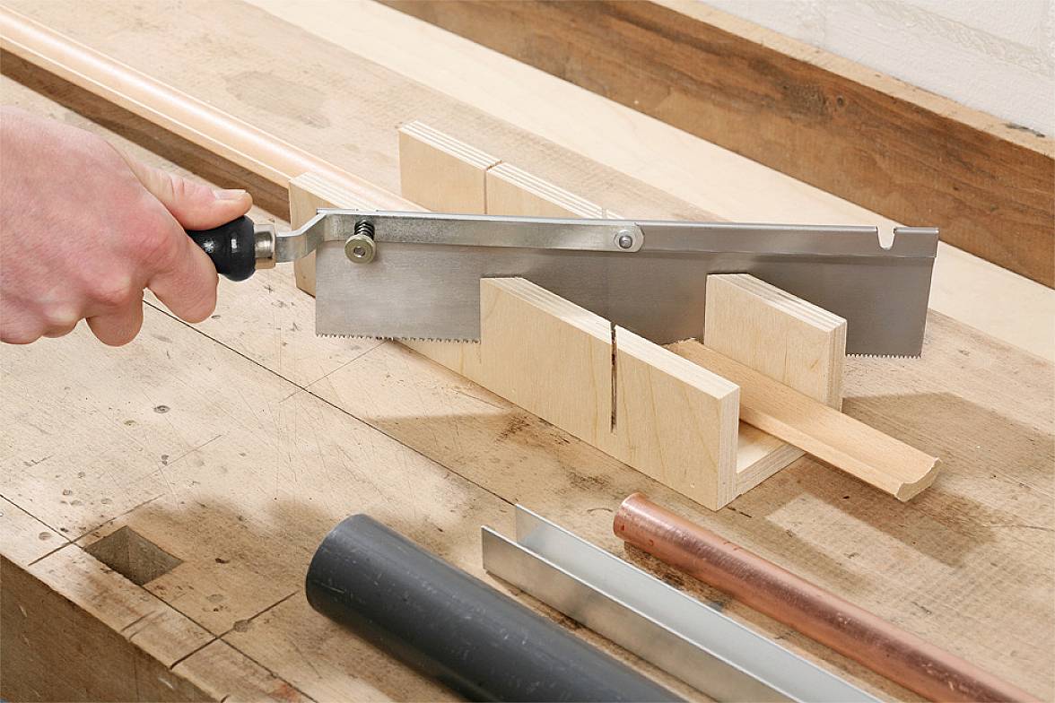 Ręka przecina kawałek drewna piłą ukośnicową na stole warsztatowym. W tle leżą kolejne narzędzia i materiały.