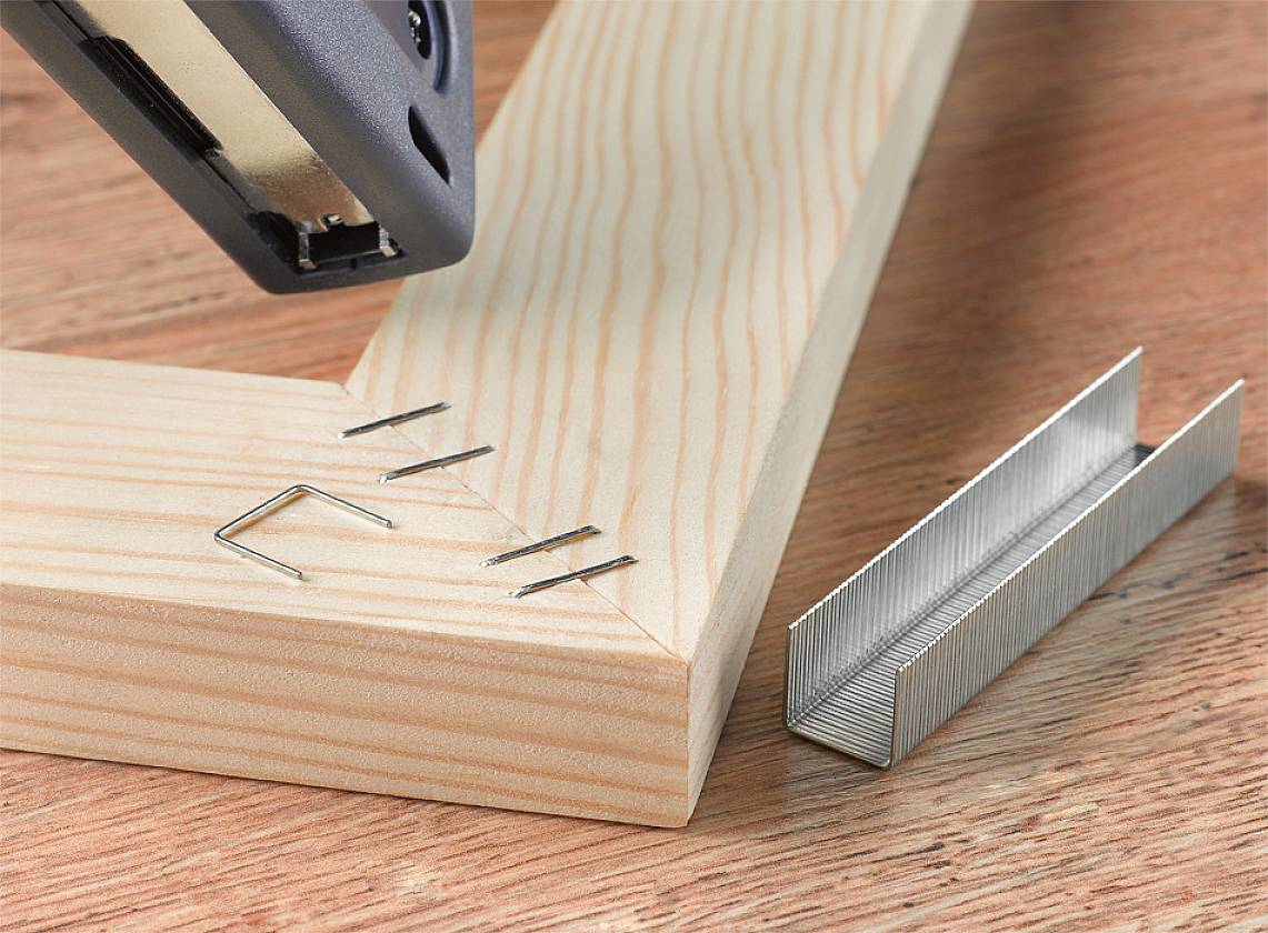 'Zszywacz mocuje dwie drewniane części pod kątem. Luźny element zszywki leży obok, na drewnianej powierzchni.'