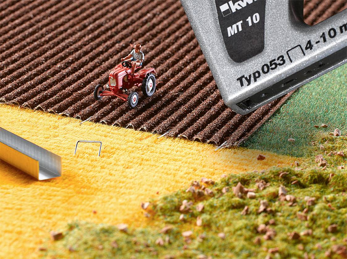 Zabawkowy traktor jedzie po strukturowanej powierzchni z filcu w ziemistych kolorach. Na pierwszym planie widoczny jest duży zszywacz.