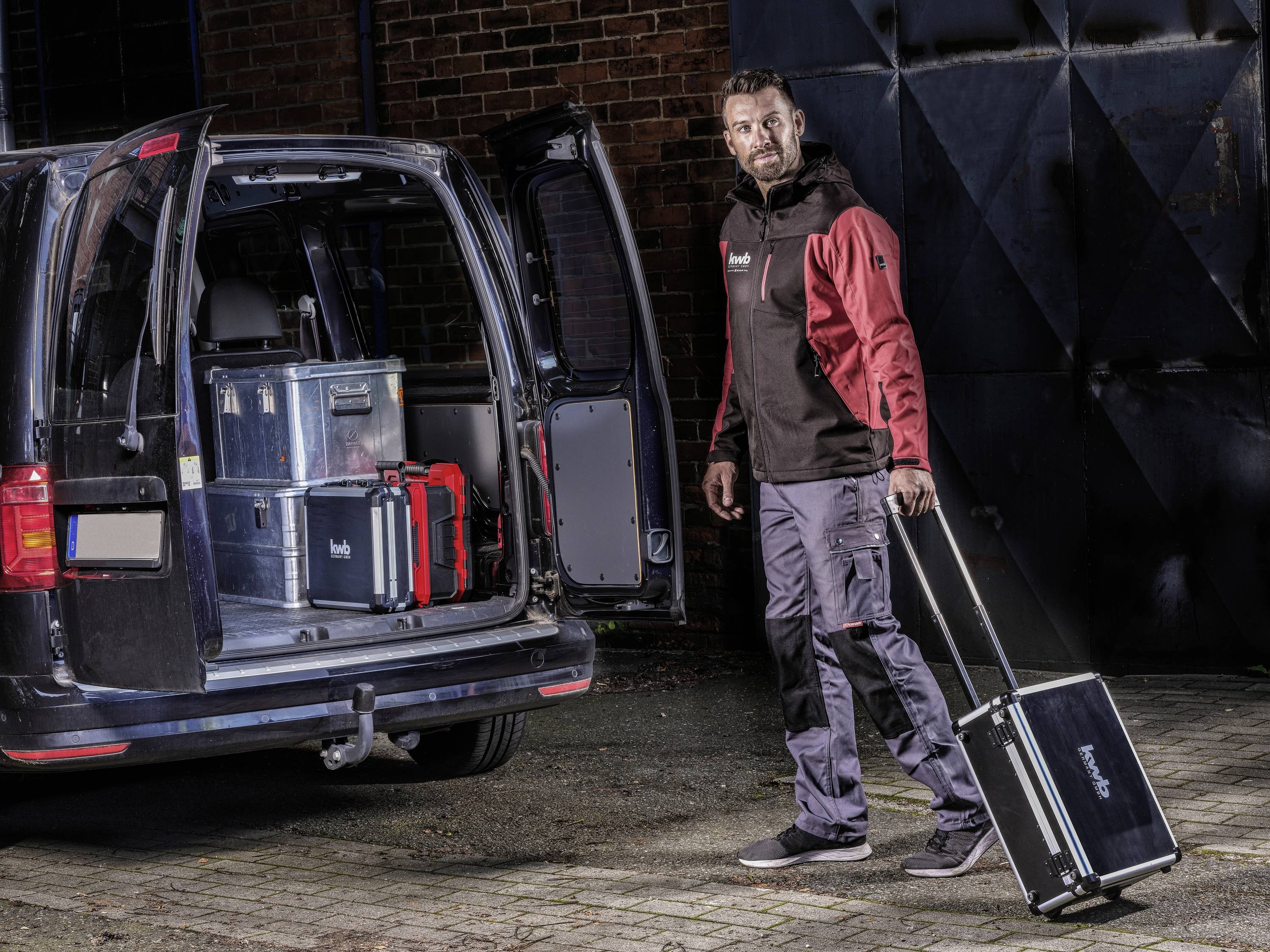 Mężczyzna ciągnie walizkę na kółkach z narzędziami przed otwartym samochodem dostawczym załadowanym kilkoma walizkami na narzędzia. Jest ciemno.