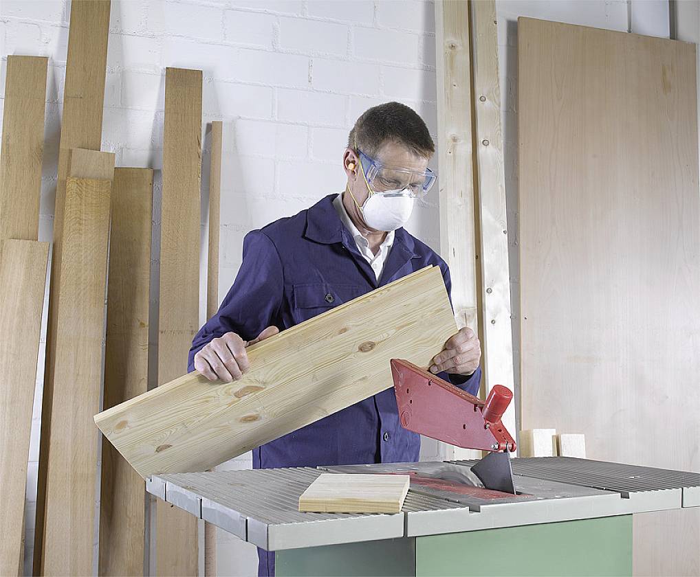 Mężczyzna w stroju ochronnym pracuje przy stołowej pile tarczowej, przecinając kawałek drewna. W tle stoją deski drewniane.