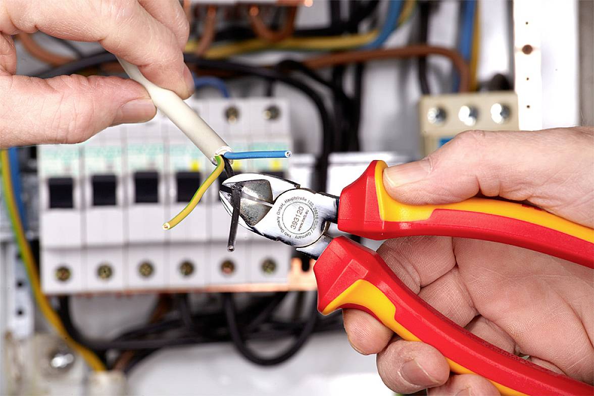 Dwie ręce trzymają obcinacz do przewodów i przecinają kabel przed elektryczną skrzynką bezpiecznikową.