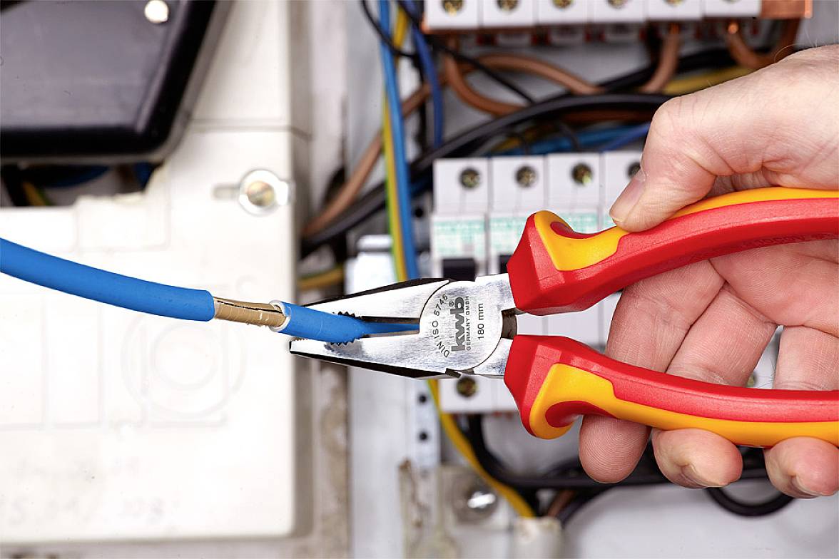 Jedna ręka trzyma obcinacz do kabli i przycina niebieski przewód w otwartej skrzynce bezpieczników elektrycznych.