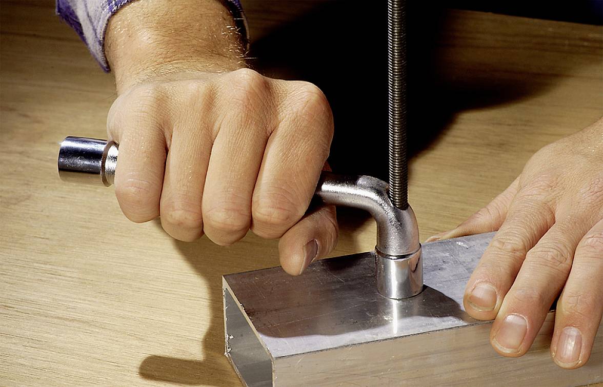 Osoba używa ścisku śrubowego, aby zamocować kawałek metalu na drewnianym stole. Dłonie mocno trzymają zacisk.