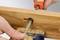 Dwie ręce pilują śrubę w desce drewnianej, przy czym powstają wióry. Jedna ręka przytrzymuje deskę, druga pilnik.
