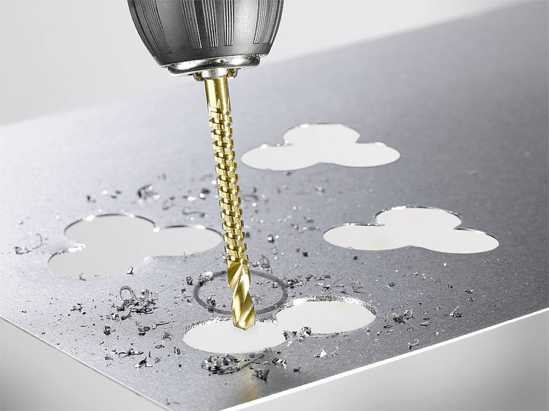 Wiertło przebija blachę metalową, tworząc wzór w kształcie koniczyny. Wokół otworów rozrzucone są wióry metalowe.