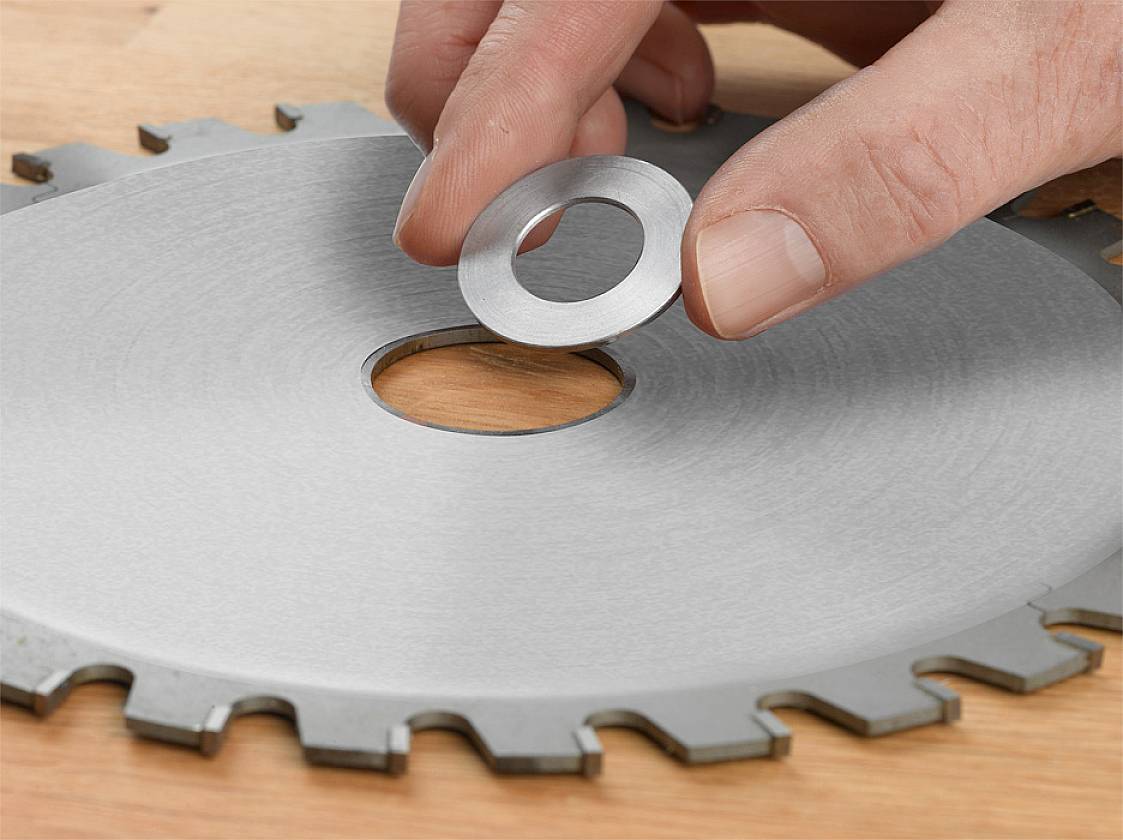 Ręka kładzie mały metalowy pierścień na otworze większej tarczy piły leżącej na stole.