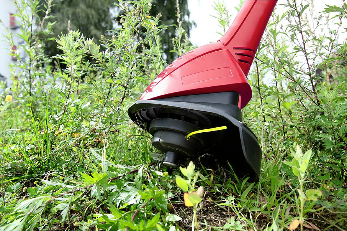 Czerwona kosiarka do trawy przycina wysoką trawę i chwasty w ogrodzie. W tle widoczne są krzewy i drzewa.