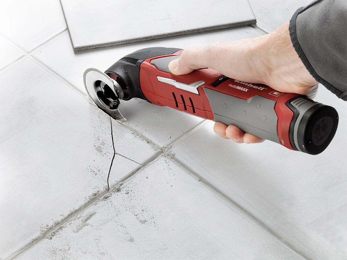 Dłoń trzyma oscylujące narzędzie z czerwonym uchwytem, przecinające płytkę wzdłuż rysy na białej podłodze ceramicznej.