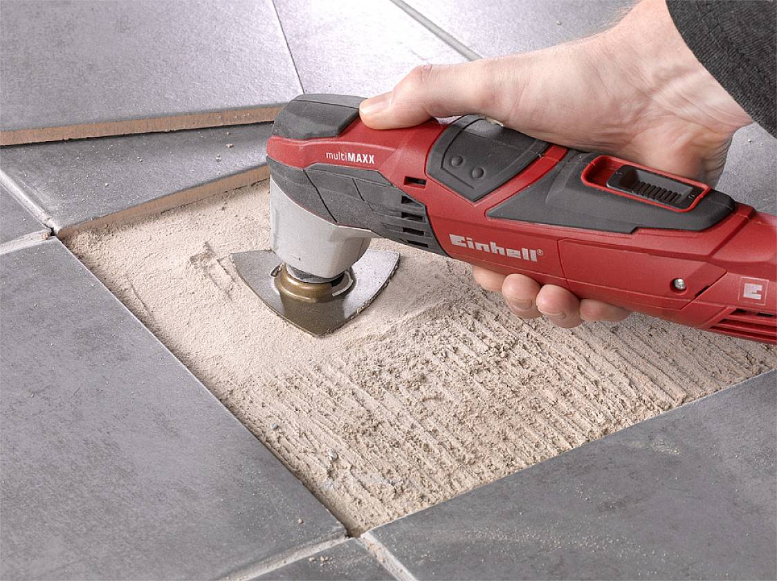 Ręka trzyma oscylacyjne narzędzie wielofunkcyjne, które usuwa fugę między szarymi płytkami.