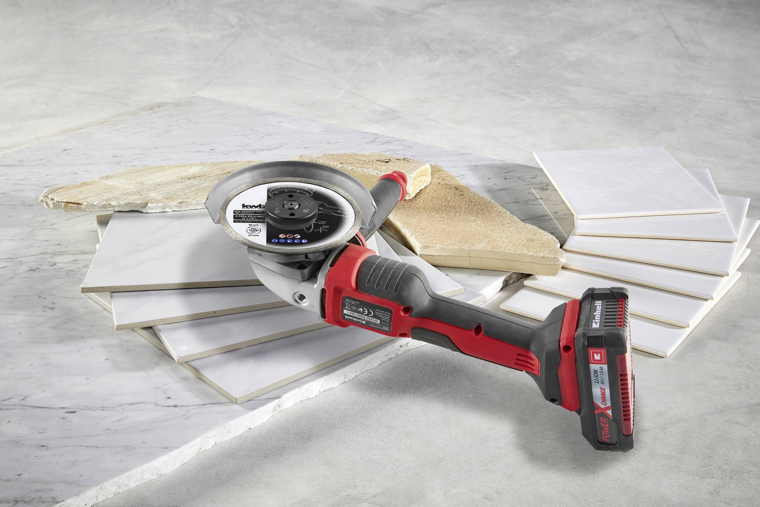 Na białych stosach płytek, na szarym podłożu, leży czerwona i czarna szlifierka kątowa.
