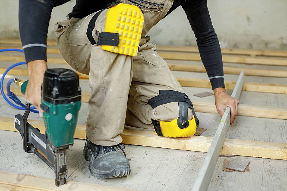 Osoba pracuje klęcząc w żółtych nakolannikach na drewnianej ramie; używa gwoździarki pneumatycznej. Prace remontowe lub budowlane.