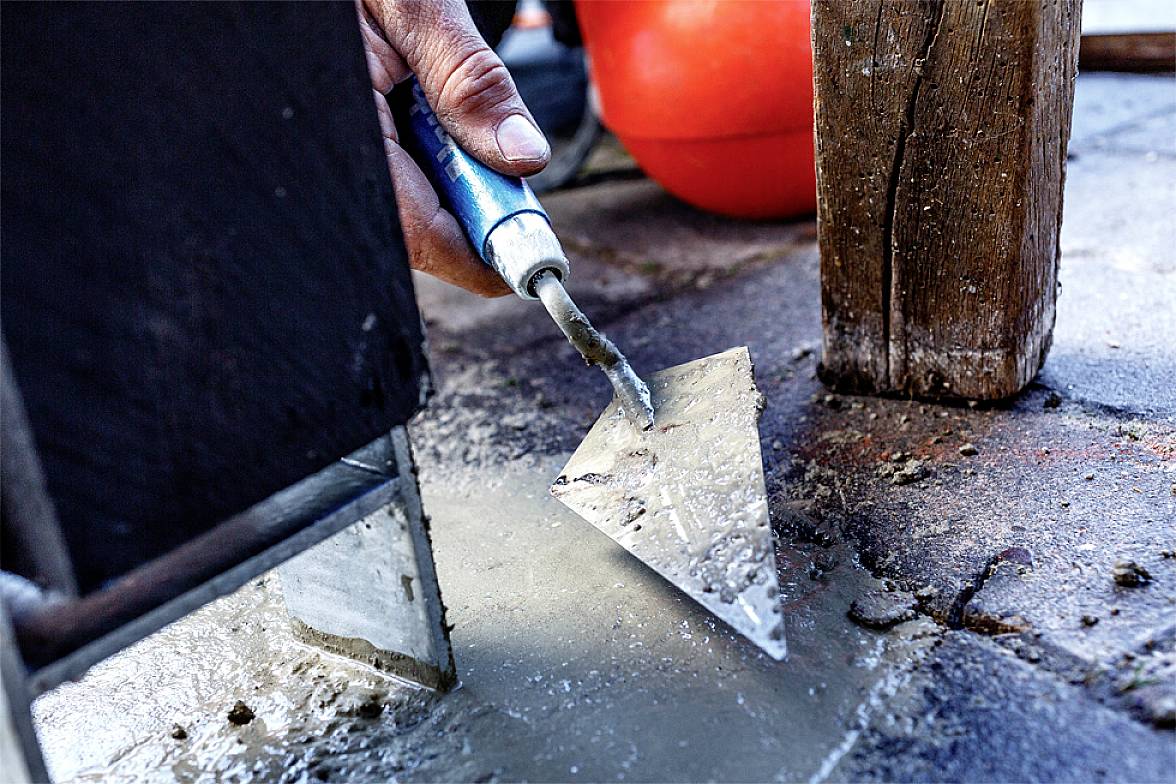 Dłoń trzyma pacę, która służy do wygładzania i kształtowania świeżego cementu między dwiema drewnianymi strukturami.