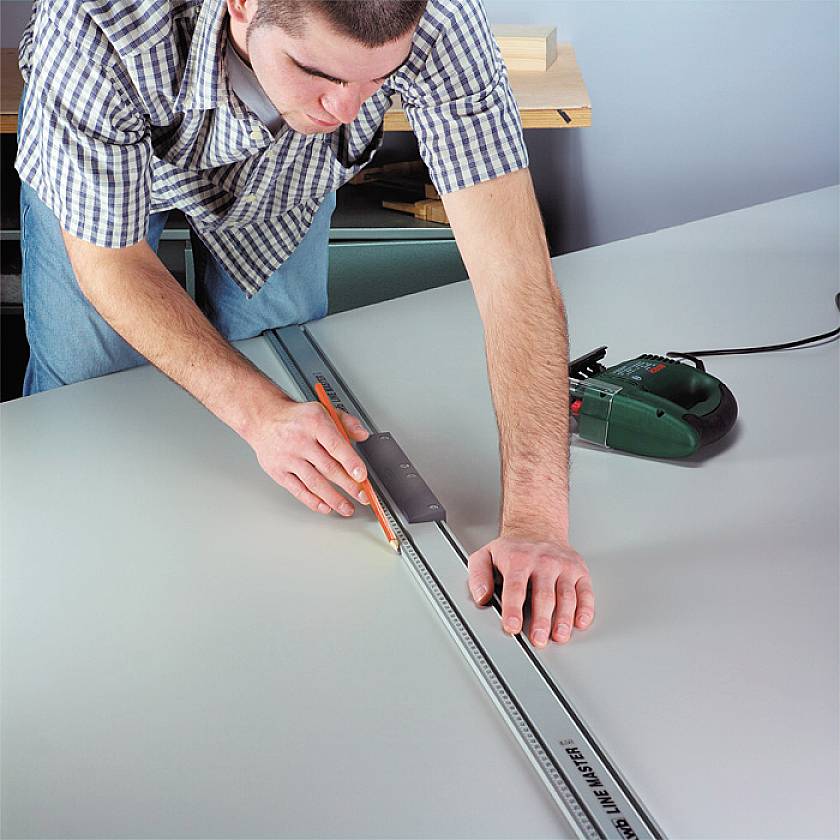 Osoba mierzy linijką na dużym stole, przygotowując się do cięcia, z elektryczną ręczną piłą tarczową w tle.