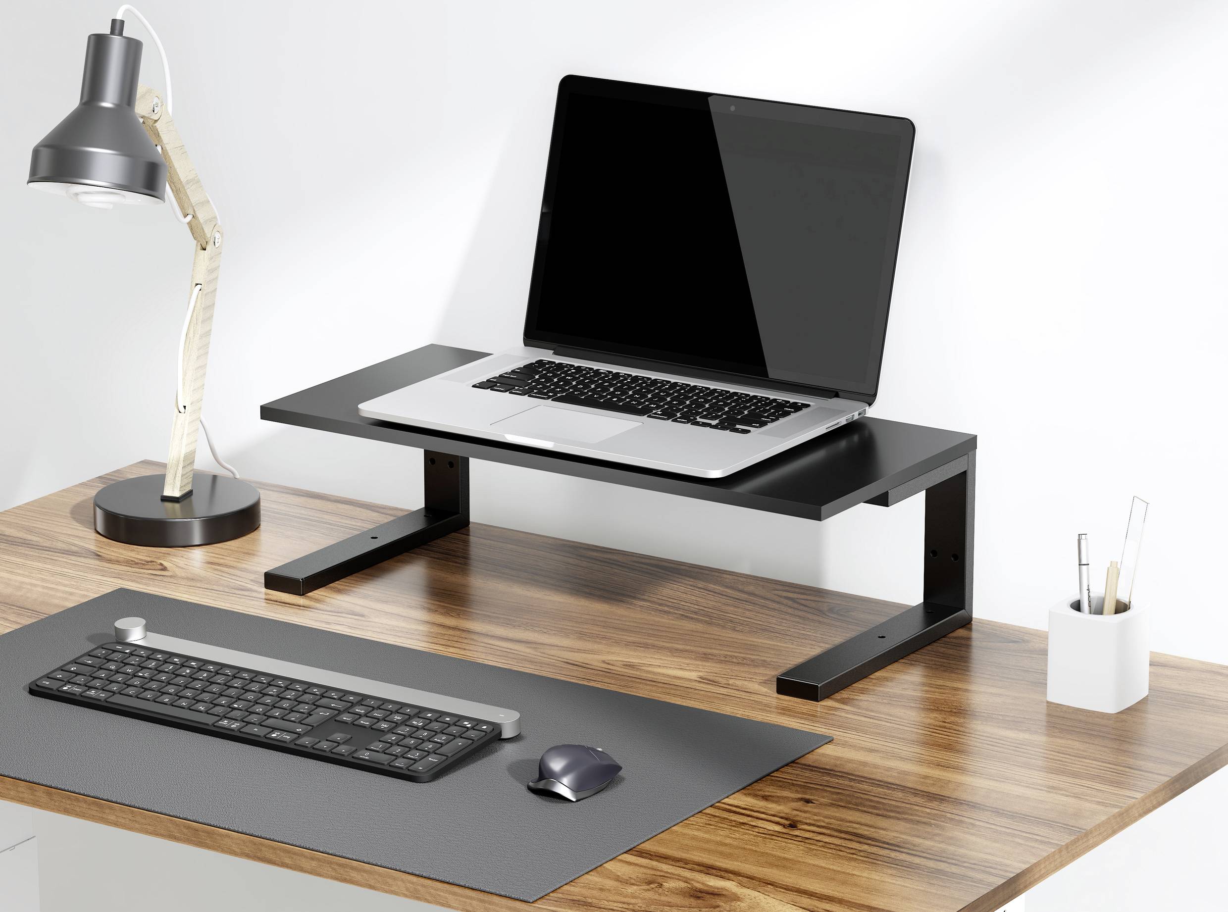 Laptop stoi na podwyższonej czarnej podstawce na drewnianym stole. Obok znajduje się lampka biurkowa i klawiatura leżąca na blacie.