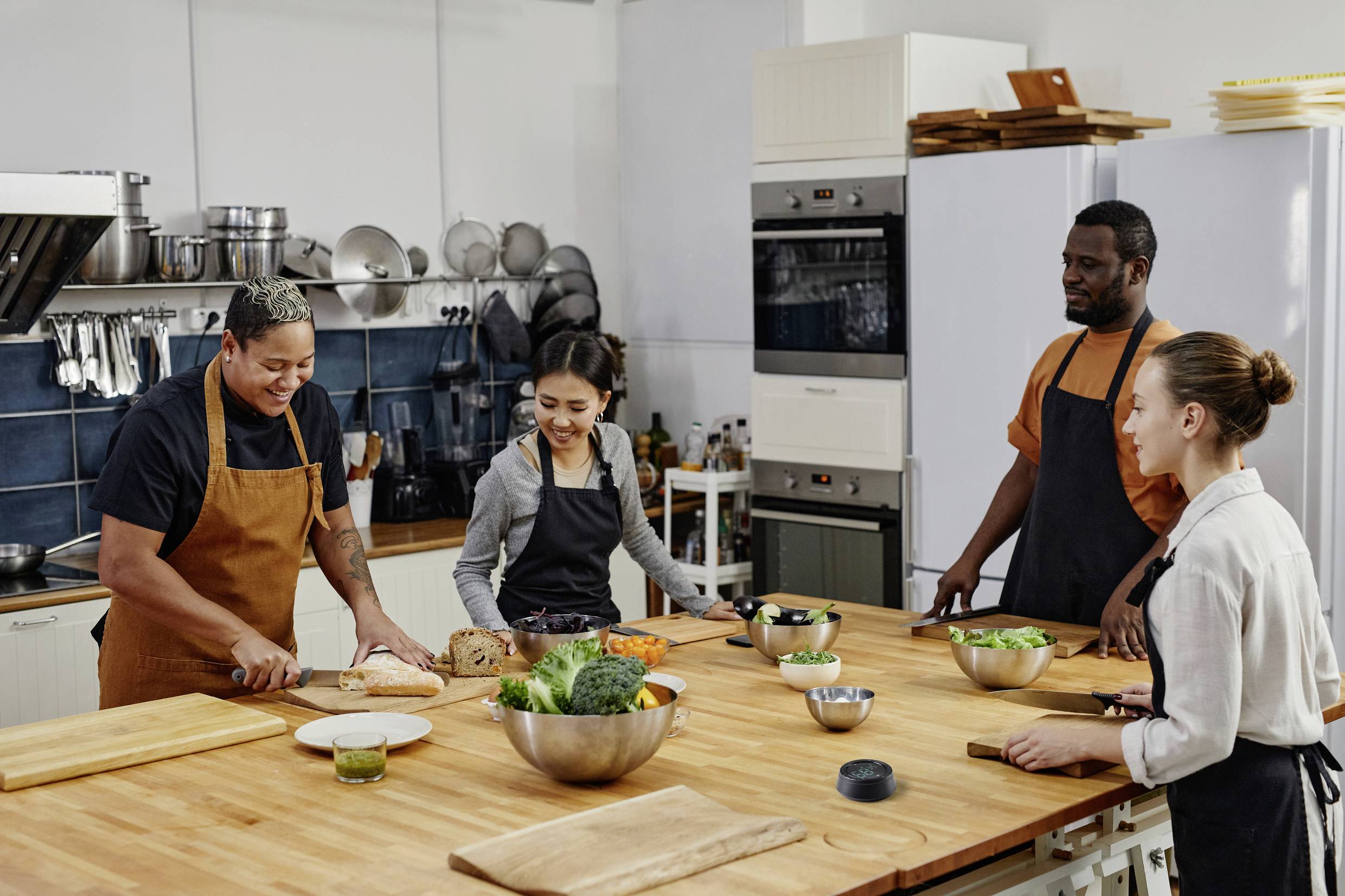 Cztery osoby w fartuchach stoją przy kuchennym stole i wspólnie przygotowują warzywa. Śmieją się i patrzą na siebie nawzajem.