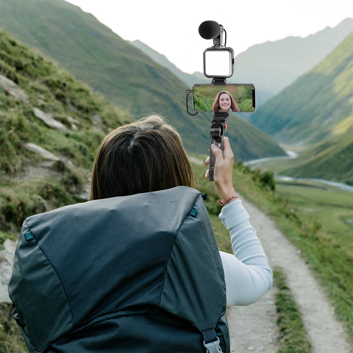 Kobieta robi sobie selfie na tle doliny górskiej. Ma na sobie duży plecak turystyczny i używa smartfona z mikrofonem i lampką.