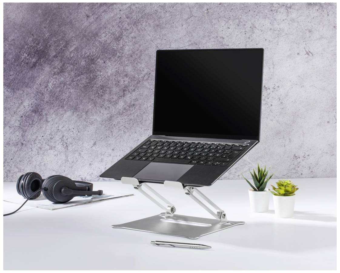 Laptop stoi na regulowanym statywie na biurku, obok słuchawek i małej rośliny.