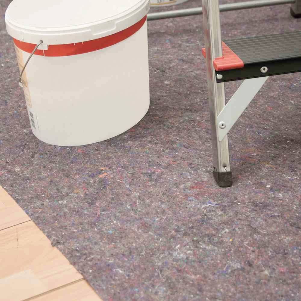 Biały wiadro z farbą stoi obok czarnej drabiny na podłodze przykrytej włókniną ochronną. Scena sugeruje prace remontowe.