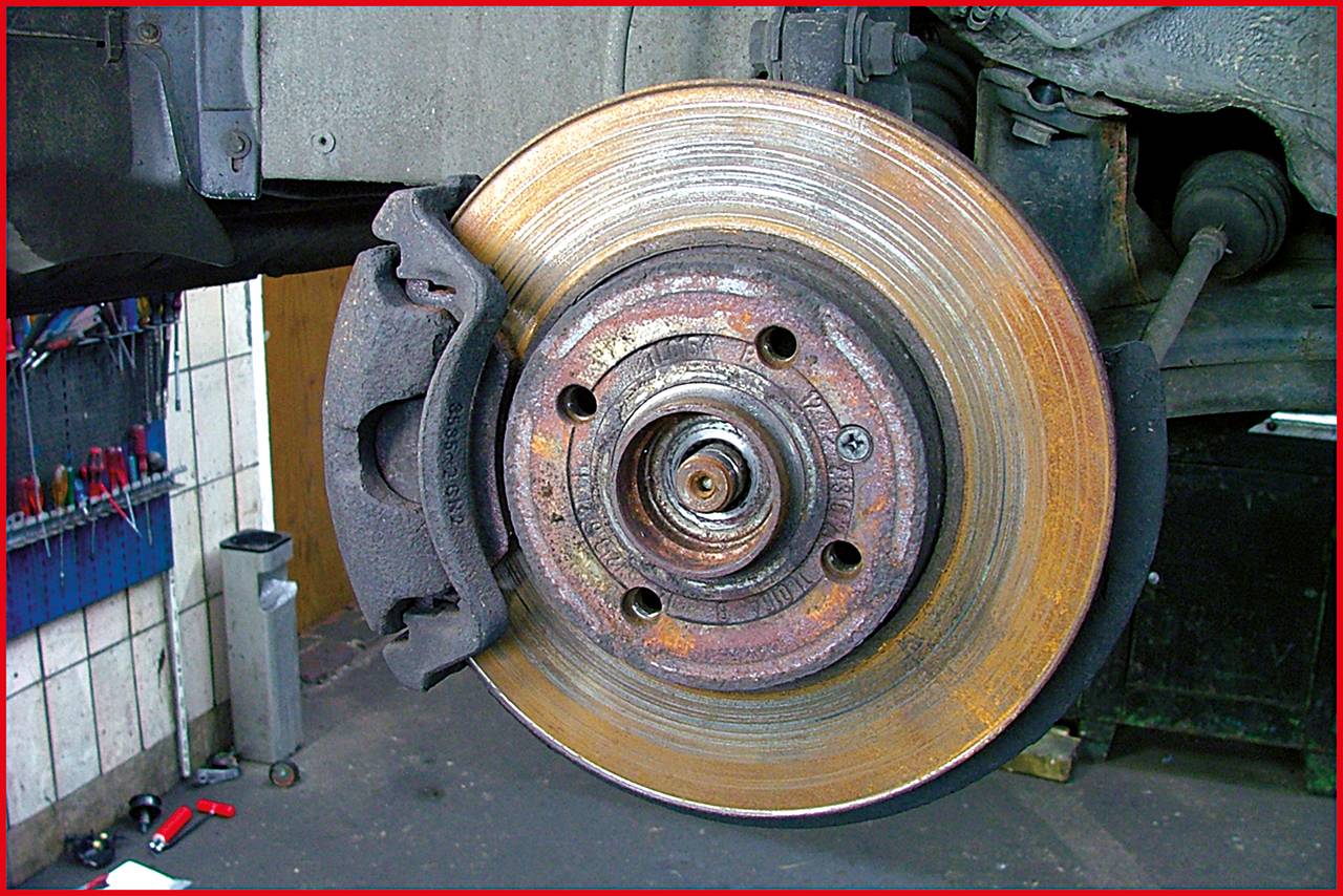 Zbliżenie na felgę samochodową z tarczą hamulcową, otoczoną klockami hamulcowymi w warunkach warsztatowych. Na tarczy widoczne są ślady rdzy.