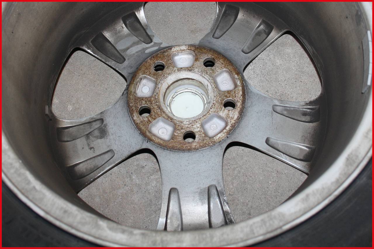 Tylna strona felgi samochodowej wykazuje ślady rdzy wokół otworów na śruby. Felga jest wykonana z metalu, a na jej wewnętrznej stronie widoczne są ciemne oznaczenia.