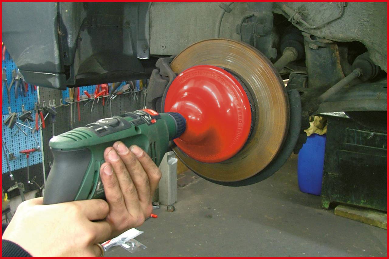 Osoba trzyma wiertarkę i obrabia zardzewiałą tarczę hamulcową w warsztacie. W tle widoczne są narzędzia.
