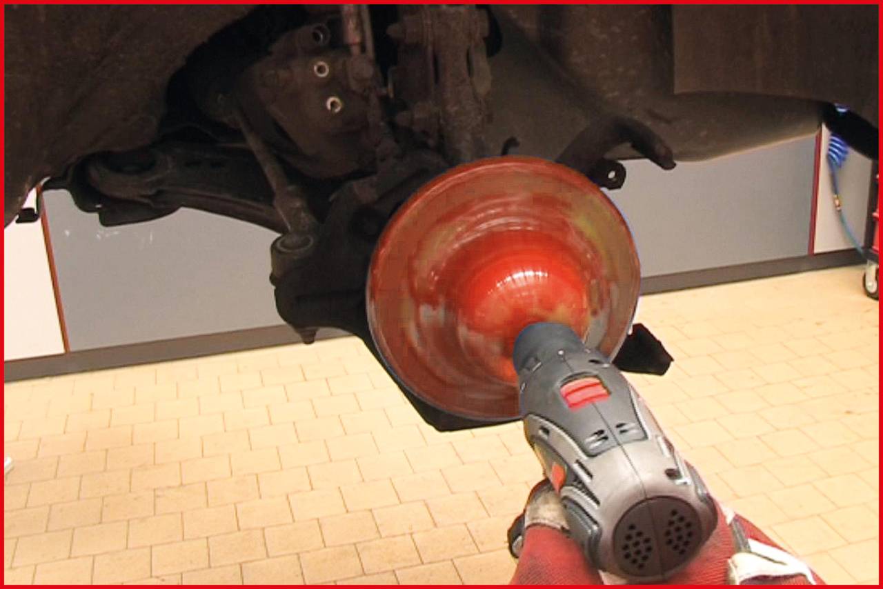 Osoba czyści układ hamulcowy samochodu elektronarzędziem w warsztacie. Na tle widoczna jest podłoga i ściany.