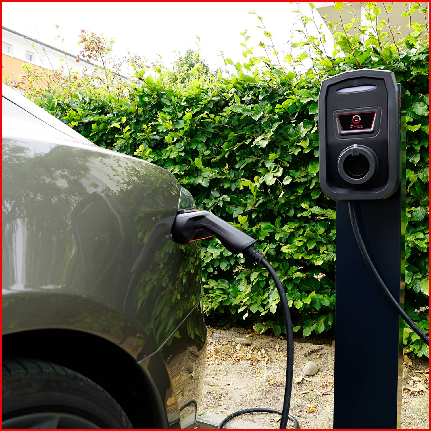 Samochód elektryczny jest ładowany przy stacji ładowania. Stacja ładowania posiada wyświetlacz i jest otoczona zielonymi krzakami.