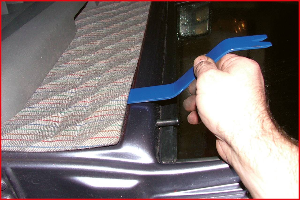 Ręka używa niebieskiego narzędzia dźwigniowego do usunięcia części samochodowej. Na desce rozdzielczej widoczna jest dywanikowa nakładka.