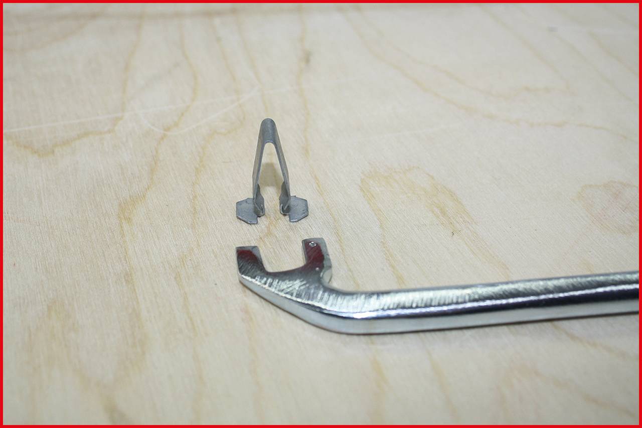 Metalowe narzędzie leży na drewnianej powierzchni, obok małego szarego elementu. Narzędzie ma zakrzywiony kształt.