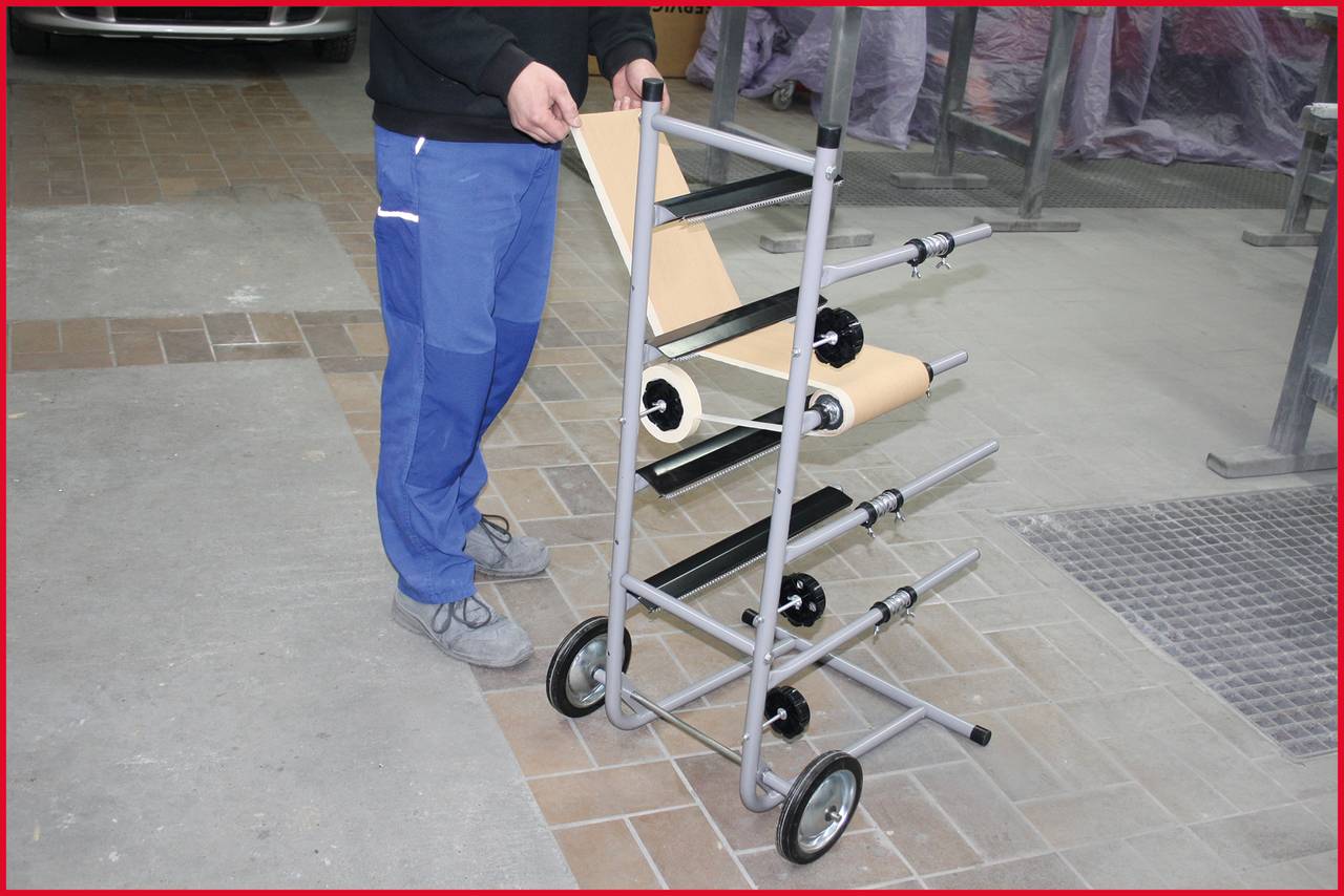 Osoba stoi obok mobilnego uchwytu na rolki papieru z kilkoma rolkami na podłodze przemysłowej lub warsztatowej.