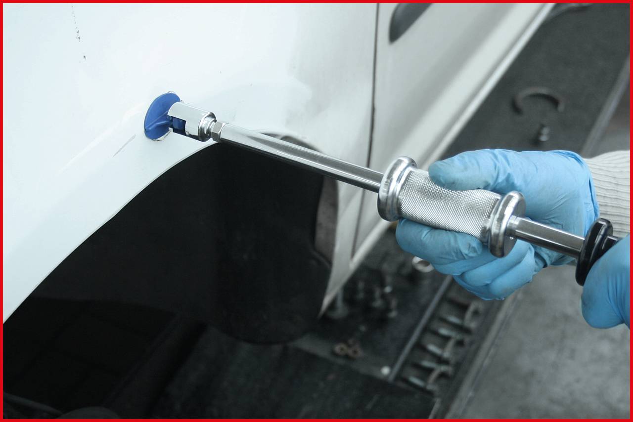 Osoba naprawia wgięcie na białym samochodzie specjalistycznym narzędziem. Ma na sobie niebieskie rękawice.