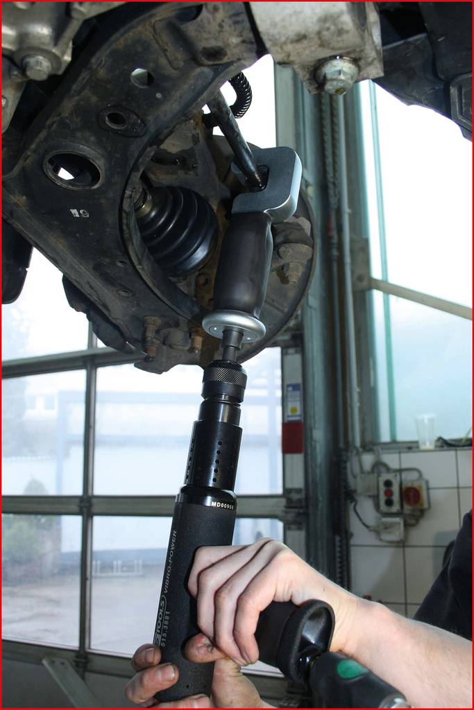 Mechanik używa specjalistycznego narzędzia do naprawy zawieszenia pojazdu. W tle widoczny jest warsztat.
