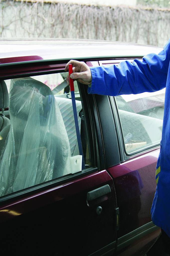 Osoba w niebieskiej kurtce otwiera drzwi bordowego samochodu za pomocą długiego narzędzia. Siedzenie jest przykryte plastikową osłoną.