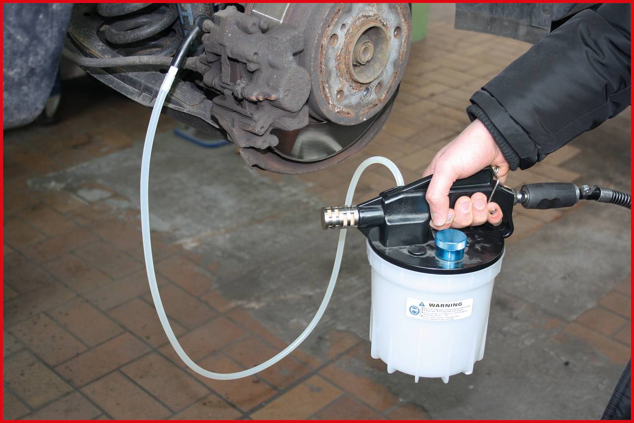Ręka trzyma urządzenie do odpowietrzania hamulców, podłączone do cylinderka hamulcowego. Podłoże przypomina warsztat samochodowy.