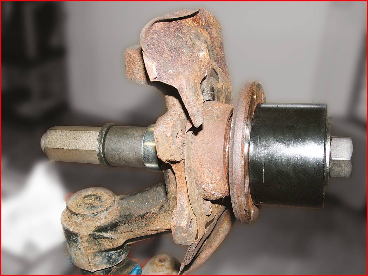 Zardzewiała metalowa część samochodowa przedstawiająca oś z piastą koła, prawdopodobnie stary lub uszkodzony element pojazdu.