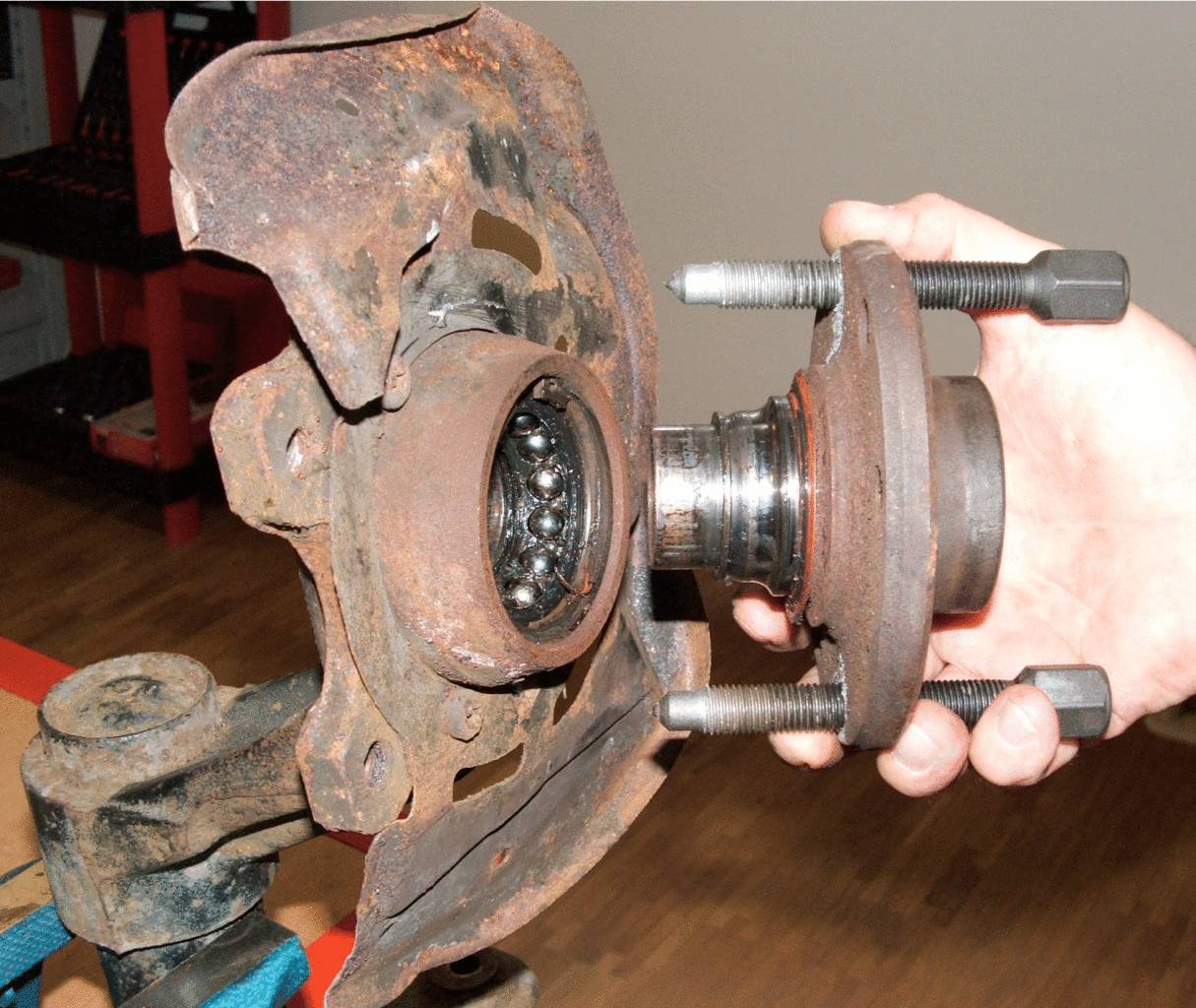 Jedna ręka trzyma zardzewiałą część samochodową z łożyskiem kulkowym, prawdopodobnie łożyskiem koła. Łożysko wydaje się zużyte i znajduje się w kontekście naprawy.