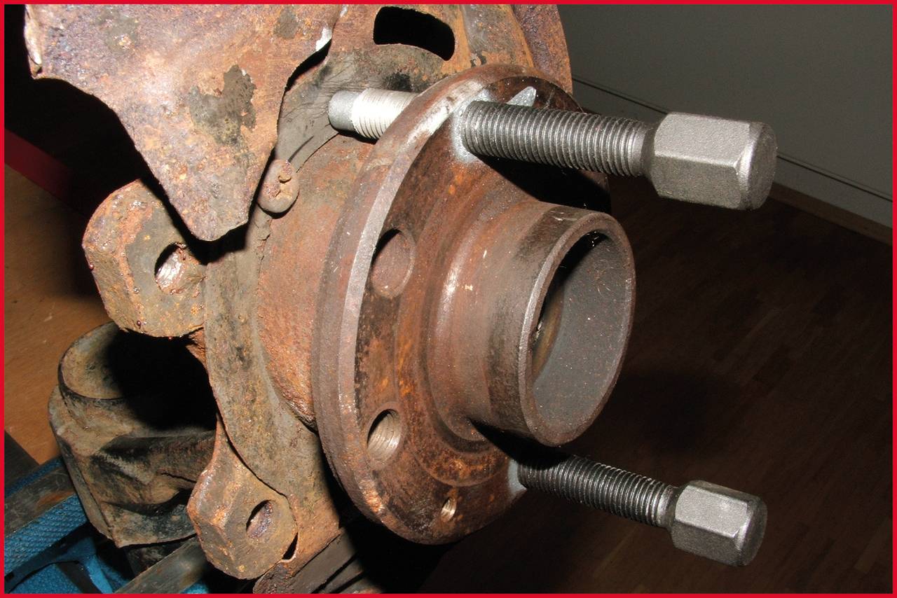 Zardzewiała część samochodowa z dwoma długimi śrubami w metalowym łożysku koła. Element leży na drewnianej powierzchni.