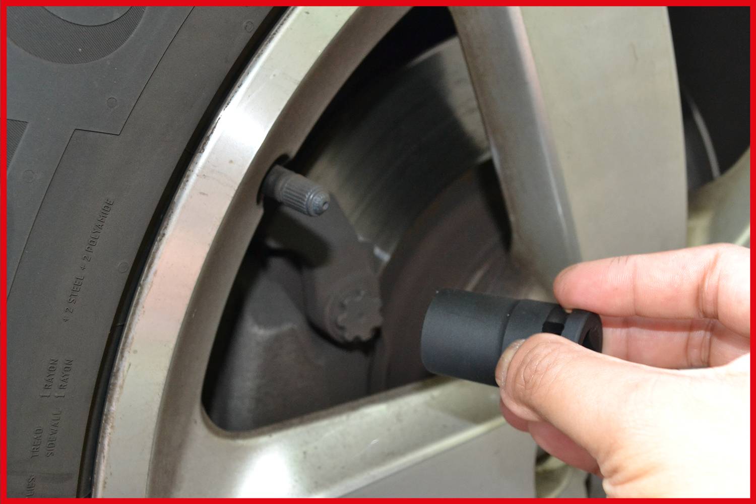 Dłoń trzyma pokrywkę zaworu magnetycznego blisko trzpienia wentyla opony samochodu, gotowa do nałożenia. Koło jest częściowo widoczne.