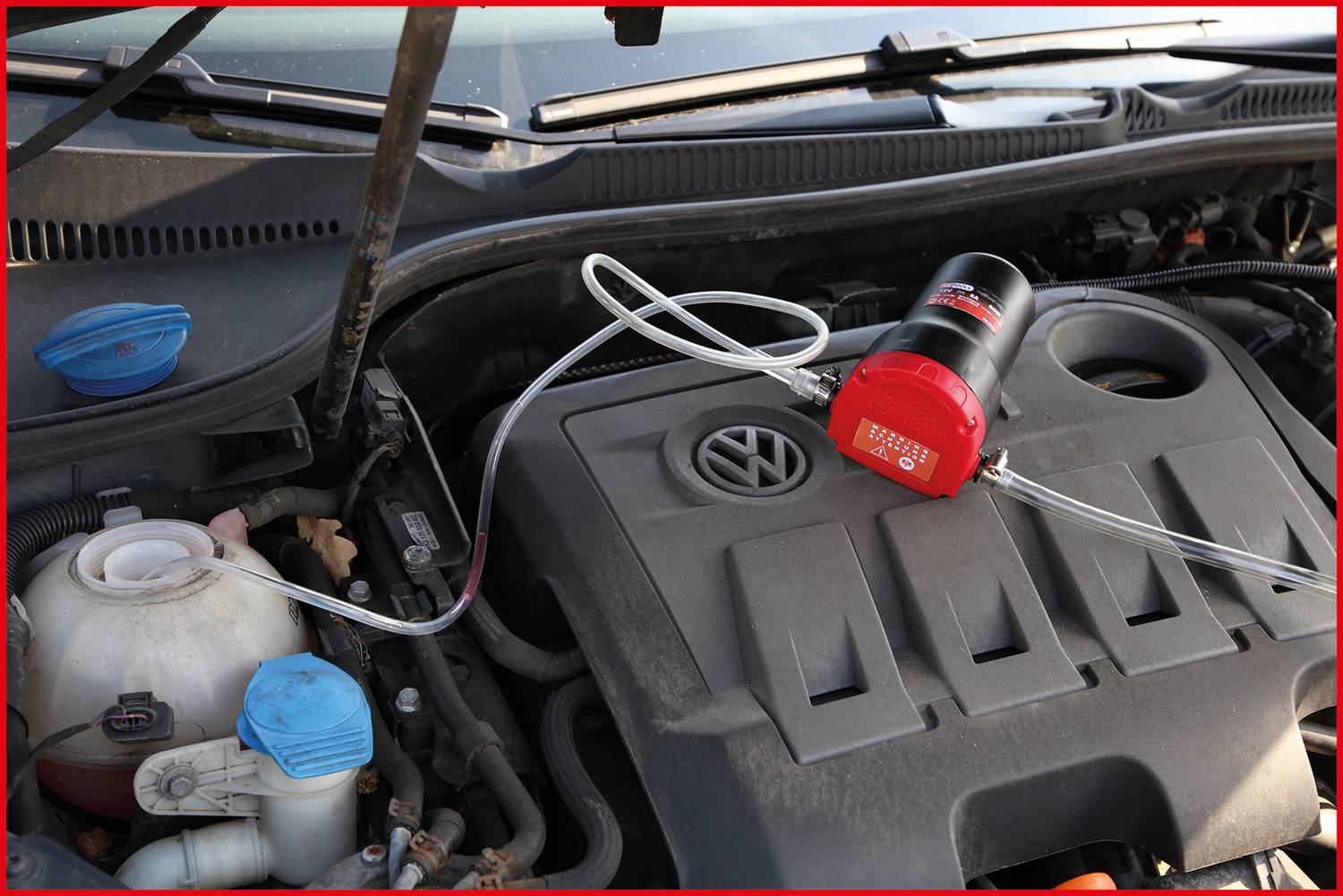 Otwarta pokrywa silnika samochodu, widać urządzenie do odsysania oleju podłączone do silnika. Po prawej stronie widoczny jest korek zbiornika płynu chłodzącego.