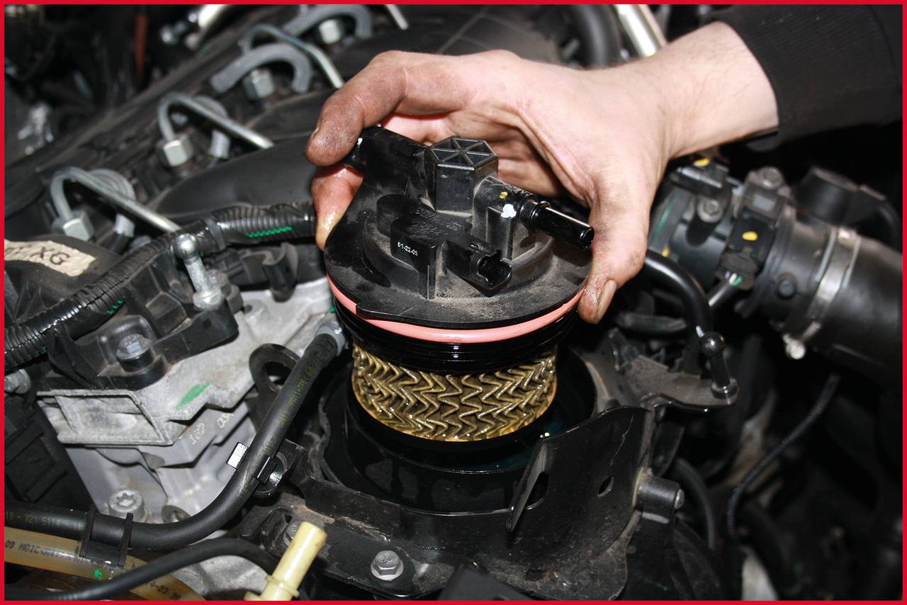 Ręka wymienia filtr oleju silnikowego w komorze silnika pojazdu. Filtr charakteryzuje się charakterystycznym zygzakowatym wzorem.