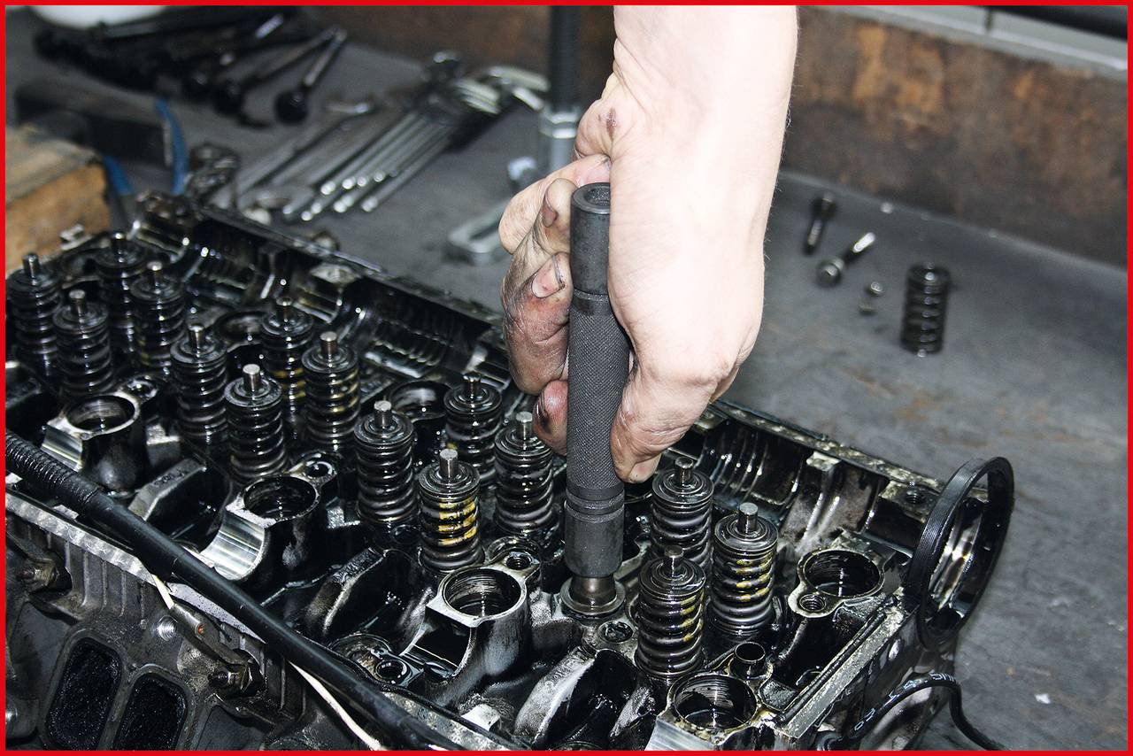 Ręka trzyma narzędzie podczas pracy przy części silnika, otoczona różnymi narzędziami samochodowymi na stole.