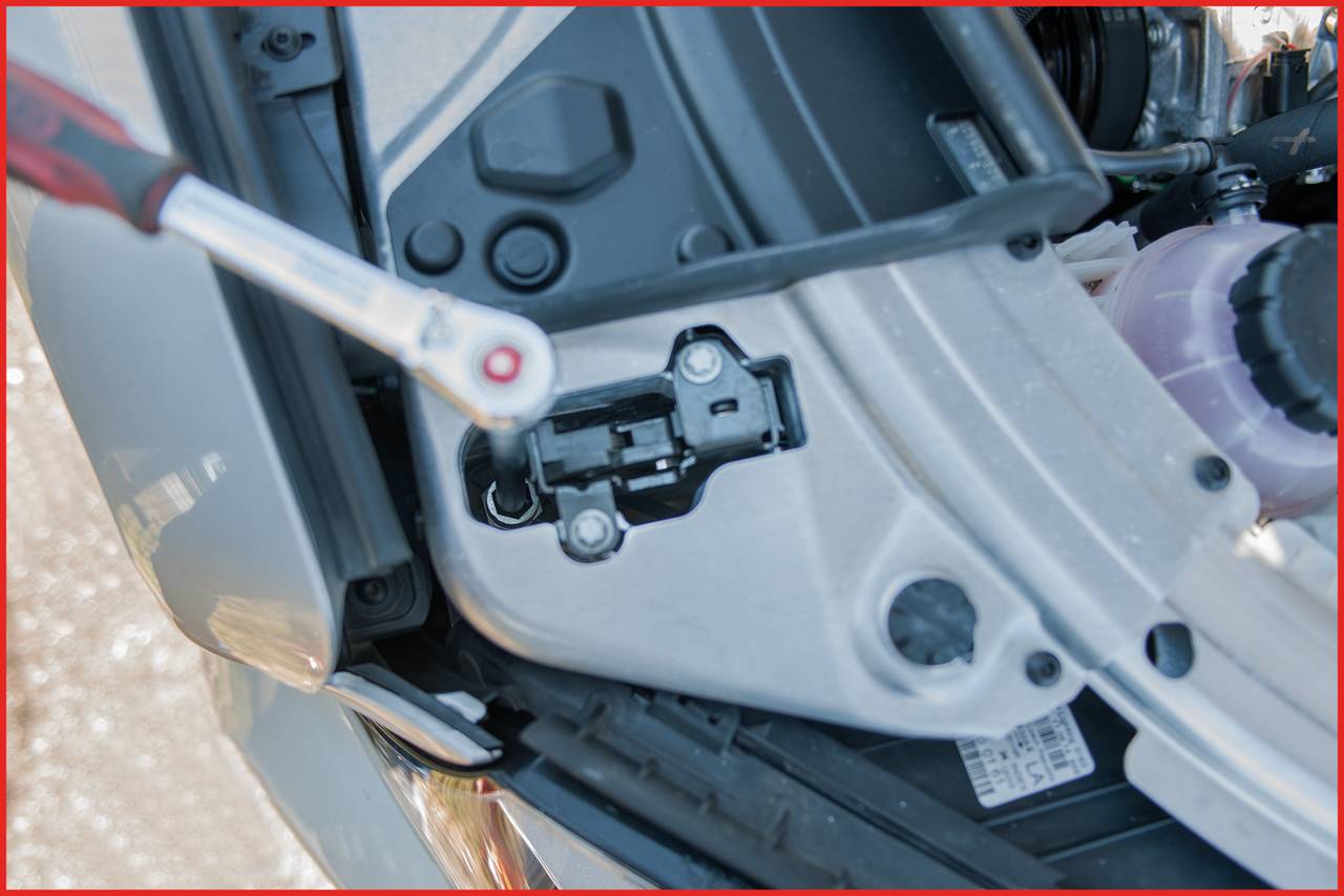 Narzędzie wskazuje na otwartą maskę silnika z widocznym zamkiem i podłączonymi przewodami.